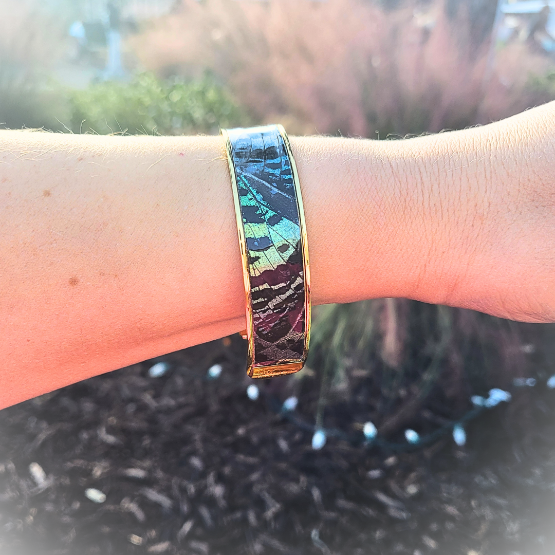 Photo of the Midnight butterfly cuff bracelet on a wrist with outdoor background. The bracelet has blue-green hues and pattern of the Madagascar Sunset Moth, with gold-plated brass hardware.